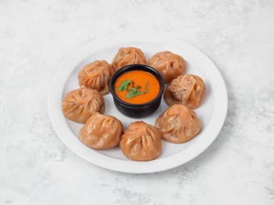 Steamed lamb momos around a bowl of orange sauce on a white plate