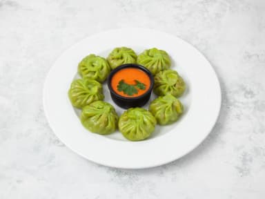 Eight green vegetable momos around a bowl of orange sauce on a white plate